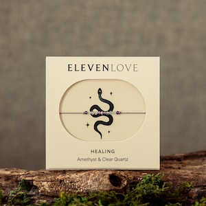 Op de afbeelding: Een beige kaart met de tekst "ELEVENLOVE" in zwarte letters. De kaart heeft een zwarte slangillustratie met sterren en een rij amethyst- en helder kwarts kralen. De tekst "HEALING" en "Amethyst & Heldere Kwarts" staat onder de illustratie.