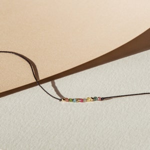 May include: A delicate necklace with a row of colorful gemstones on a black cord. The gemstones are a mix of green, pink, and yellow.