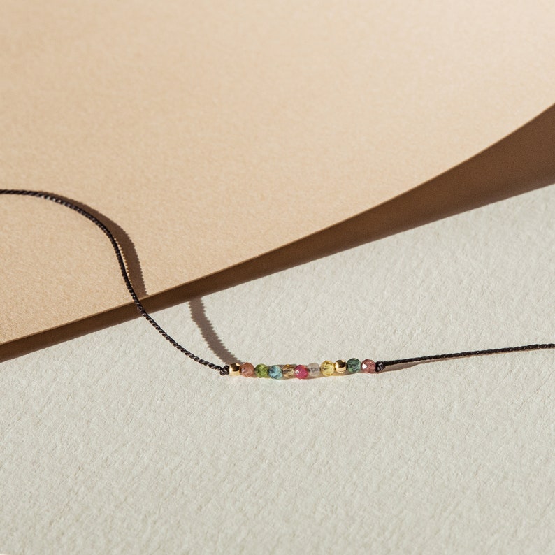 May include: A delicate necklace with a row of colorful gemstones on a black cord. The gemstones are a mix of green, pink, and yellow.