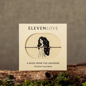 May include: A white card with the text "ELEVENLOVE" in black lettering. The card features a black and white illustration of a woman with stars in her hair. The card also includes the text "A WISH FROM THE UNIVERSE" and "Rainbow Tourmaline".