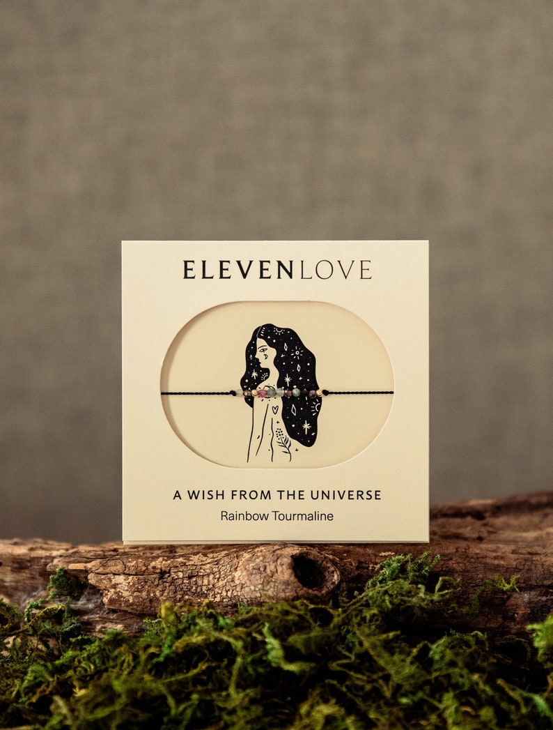 May include: A white card with the text "ELEVENLOVE" in black lettering. The card features a black and white illustration of a woman with stars in her hair. The card also includes the text "A WISH FROM THE UNIVERSE" and "Rainbow Tourmaline".