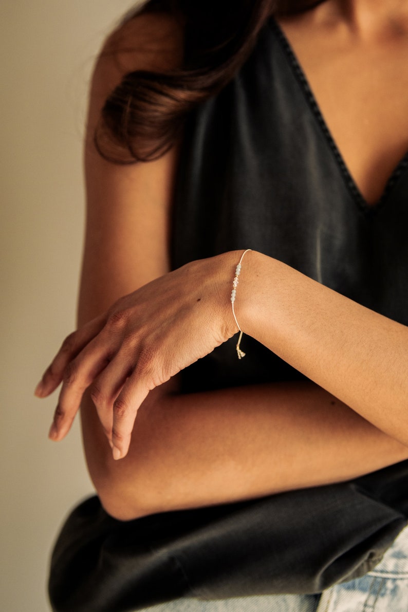 Peut inclure: Un bracelet d&eacute;licat avec de petites pierres gris clair sur une fine cordelette blanche. Le bracelet est nou&eacute; avec un simple n&oelig;ud.