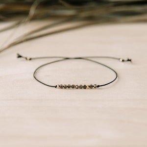 May include: A delicate black string bracelet with a row of small, dark brown faceted beads and gold accents.