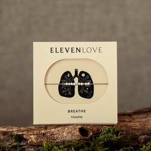May include: A white card with the text "ELEVENLOVE" and "BREATHE Howlite" on it. The card features a black and white illustration of lungs with a moon, sun, and stars inside. A white and pink howlite beaded bracelet is shown on the card.