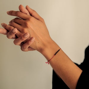 May include: A red string bracelet with small black beads. The bracelet is worn on a person's wrist, with the string tied in a knot. The background is a neutral colour.