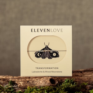 May include: A white card with the text "ELEVENLOVE" and "TRANSFORMATION" printed on it. The card features a black and white illustration of a moth with a string of beads around its body. The text "Labradorite & Mixed Moonstone" is printed below the illustration.