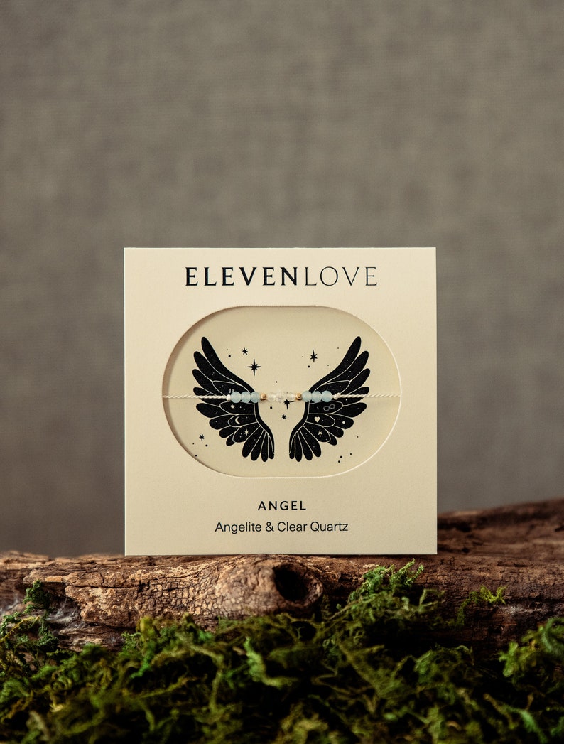 Peut inclure: Une carte blanche avec du texte noir qui dit "ELEVENLOVE" et une illustration en noir et blanc d'ailes d'ange avec des &eacute;toiles. Le texte sous l'illustration dit "ANGEL" et "Angelite & Clear Quartz".