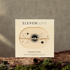 May include: A gold bracelet with an eye-shaped centrepiece, presented on a cream card. The card reads "ELEVENLOVE" and "CONNECTION", with material details: Pearl, Pink Opal, and Clear Quartz. The bracelet is displayed against a natural backdrop.