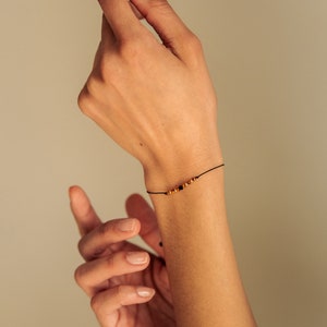 Puede incluir: Una muñeca con una pulsera simple de cuerda negra con pequeñas cuentas doradas y negras.