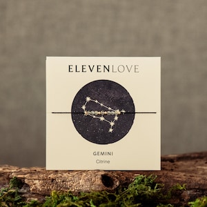 Könnte beinhalten: Eine Karte mit dem Text "ELEVENLOVE" und einem schwarzen Kreis mit einem goldenen Sternbild-Design. Der Text "GEMINI Citrine" befindet sich unter dem Kreis.