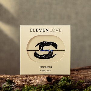 Peut inclure: Une carte blanche avec le texte "ELEVENLOVE" et "EMPOWER Lapis Lazuli". La carte présente une illustration en noir et blanc de deux mains tenant un bracelet en cordon avec des perles bleues.