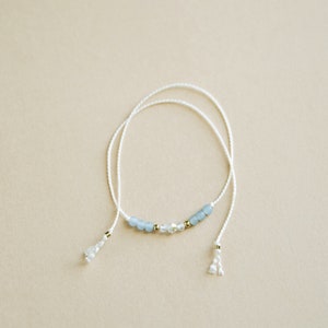 Peut inclure: Un bracelet d&eacute;licat en cordon blanc avec des perles de verre bleu clair et transparentes et des accents dor&eacute;s. Le bracelet a deux glands aux extr&eacute;mit&eacute;s.