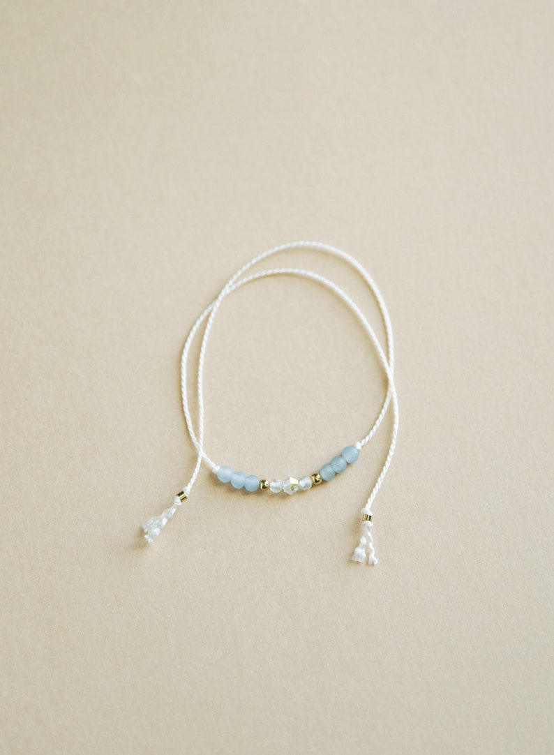 Peut inclure: Un bracelet d&eacute;licat en cordon blanc avec des perles de verre bleu clair et transparentes et des accents dor&eacute;s. Le bracelet a deux glands aux extr&eacute;mit&eacute;s.