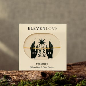 Puede incluir: Una tarjeta color crema con el texto "ELEVENLOVE" y una ilustración de manos, ojos y una estrella. La tarjeta dice "PRESENCE" y "Yellow Opal & Clear Quartz". Se muestra una pulsera dorada con cuentas amarillas.
