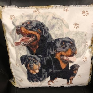Op de afbeelding: Een vierkant sierkussen met een geweven tapijtdesign van drie zwart-bruine Rottweilers. De achtergrond is gebroken wit met verspreide bruine pootafdrukken en een afwerking met gerafelde randen aan alle vier de zijden van de hoes.