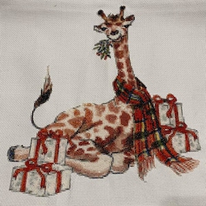 May include: A cross-stitch pattern of a giraffe wearing a red and black plaid scarf and holding a sprig of holly. The giraffe is sitting on a pile of wrapped presents with red bows.