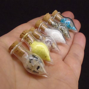 May include: A set of six small glass bottles with cork stoppers, each filled with different coloured and textured materials. The bottles are arranged in a row on a hand, with the stoppers facing the viewer.