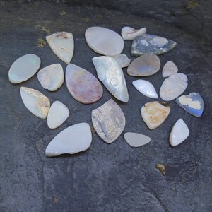 May include: A collection of 17 white, gray, and brown opal cabochons of various shapes and sizes. The cabochons are arranged on a dark gray surface.