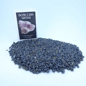 May include: A small pile of dark gray, metallic-looking gravel. A white card with the text "Scottish Gems" and "A precious piece of Scotland" is visible in the background.