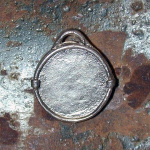 May include: A silver-colored metal pendant with a round, blank surface. The pendant has a loop at the top for attaching to a chain or cord.