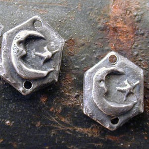May include: Two silver metal charms in the shape of a crescent moon with a star. The charms have a hexagonal shape with three holes on each side.