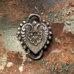 May include: A silver heart-shaped charm with intricate details and a loop at the top. The charm is on a rusty metal surface.