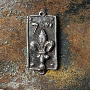 May include: A rectangular, silver-toned pendant featuring a fleur-de-lis design. The pendant has the numbers "7 AW" etched at the top. The pendant has a loop at the top for hanging.