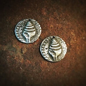 May include: Two silver-toned metal charms with a raised design of a seashell. The charms have a textured, slightly rough surface.