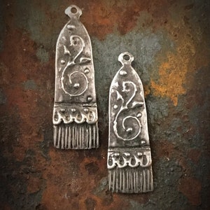 May include: Two silver metal pendants with a decorative floral design and a fringe detail. The pendants are on a rusty brown background.