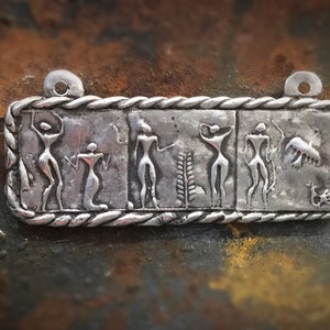 May include: A silver pendant with a rectangular shape and a rope-like border. The pendant features five figures in a stylised design, possibly representing dancers or figures in a ritualistic scene. The pendant has two holes at the top for attaching a chain or cord.