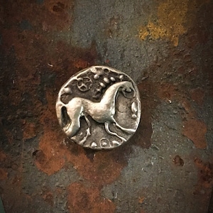 May include: A small, circular, silver coin with a detailed relief of a horse. The horse is shown in profile, with a flowing mane and tail. The coin has an aged, antique appearance, with intricate details around the horse.