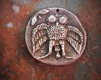 May include: A silver pendant featuring a detailed owl with large eyes and outstretched wings. The owl is surrounded by a circular frame with a textured surface.