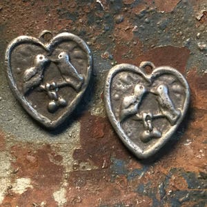 May include: Two silver heart-shaped charms with a pair of birds perched on a branch inside each heart. The birds are facing each other and appear to be kissing.