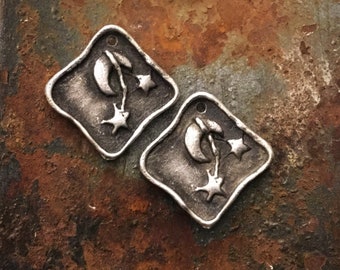May include: Two silver-toned metal charms shaped like a square with rounded corners. Each charm features a crescent moon and two stars on a stem.