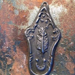 May include: A silver-coloured metal pendant with an ornate design featuring a stylised feather and a horseshoe. The pendant has a hole at the top for hanging.