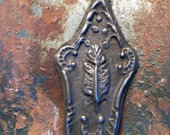Puede incluir: Un colgante de metal plateado con un diseño ornamentado que presenta una pluma estilizada y una herradura. El colgante tiene un agujero en la parte superior para colgar.