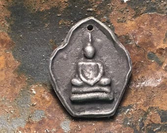May include: A silver-colored pendant featuring a detailed depiction of a seated Buddha. The pendant has a hole at the top for hanging.