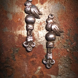 May include: Two silver bird charms with a round bead and a three-pronged base. The charms are on a rusty brown background.