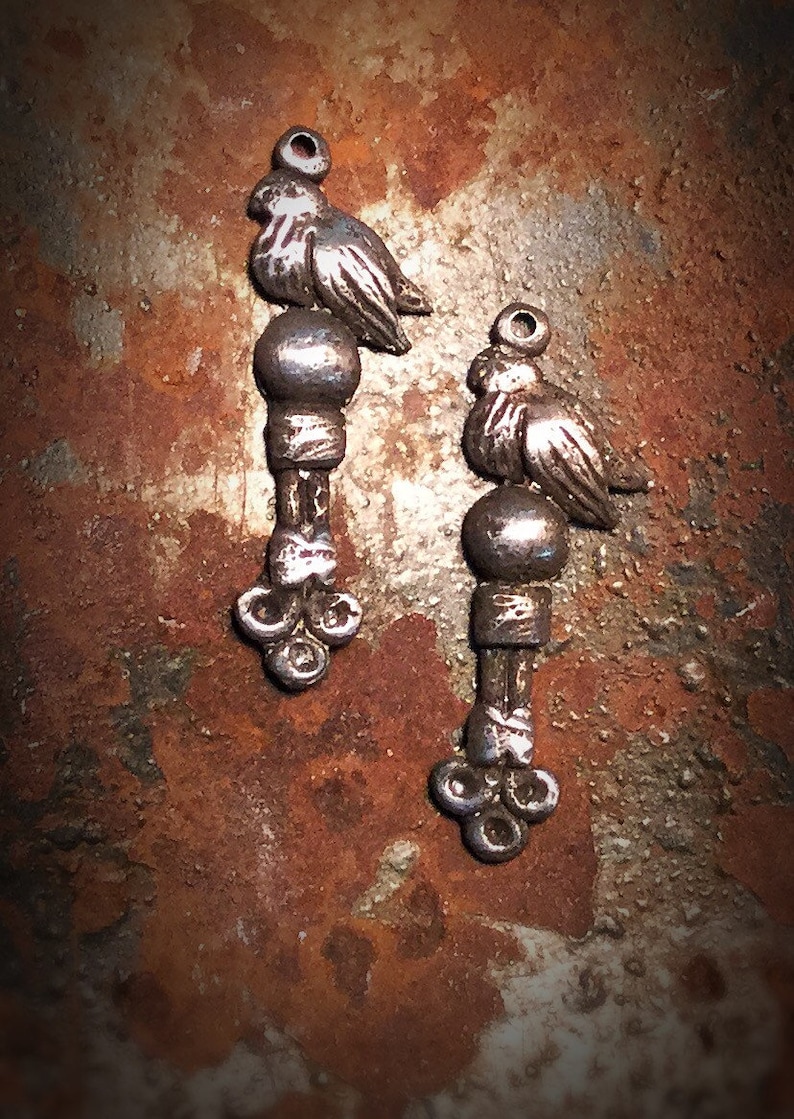 May include: Two silver bird charms with a round bead and a three-pronged base. The charms are on a rusty brown background.