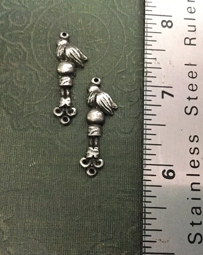 May include: Two silver-toned metal charms shaped like birds with a small ball and a decorative design at the bottom. The charms are about 1 inch long and are shown next to a ruler.