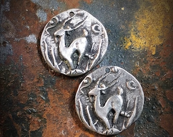 May include: Two silver metal pendants with a deer design. The deer are facing to the right and are in a stylized, almost abstract design. The pendants are round and have a slightly textured surface.