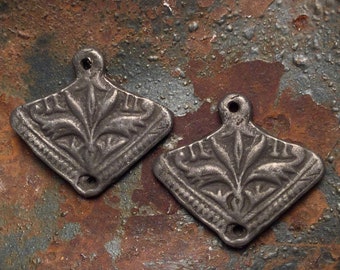 May include: Two antique silver-toned metal pendants. Each pendant is diamond-shaped with a floral design in the center and a small hole at the top for hanging. The pendants have a distressed, aged appearance.