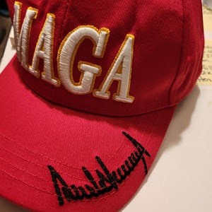 May include: Red baseball cap with white and gold embroidered text "MAGA" and black embroidered text "Donald Trump" on the brim.