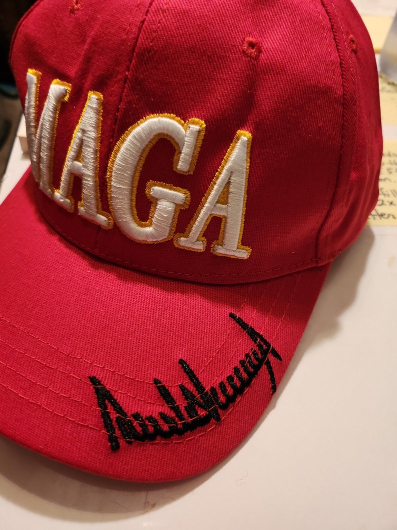 May include: Red baseball cap with white and gold embroidered text "MAGA" and black embroidered text "Donald Trump" on the brim.
