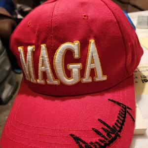 May include: Red baseball cap with a white and gold embroidered "MAGA" on the front and a black embroidered signature on the brim.