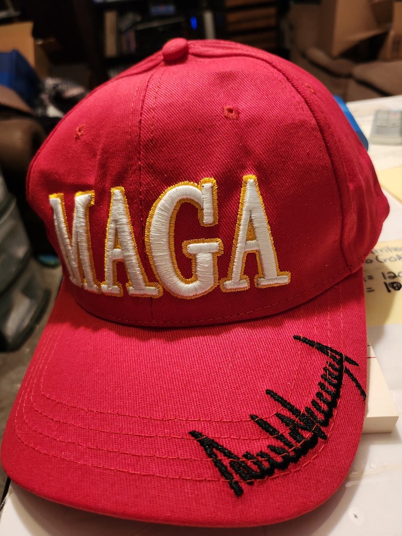 May include: Red baseball cap with a white and gold embroidered "MAGA" on the front and a black embroidered signature on the brim.