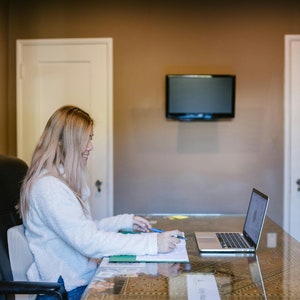 Puede incluir: Una mujer está sentada en un escritorio de vidrio en una oficina en casa, trabajando en una computadora portátil. Lleva un suéter blanco y tiene el pelo rubio. Hay un televisor en la pared detrás de ella.