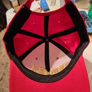 May include: Red baseball cap with a black brim and a black star design on the inside. The cap has a yellow embroidered word "MAGA" on the inside.
