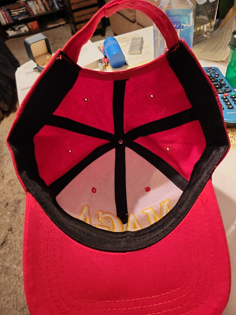 May include: Red baseball cap with a black brim and a black star design on the inside. The cap has a yellow embroidered word "MAGA" on the inside.