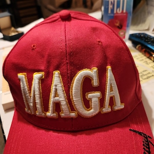 May include: Red baseball cap with a white and gold embroidered "MAGA" logo. The brim of the cap has a black embroidered design.